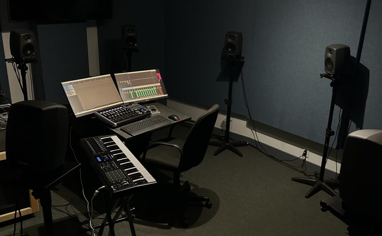 MCS Far A darkly lit room with acoustic treatment on the walls, containing a desk with two computer monitors, a midi controller, and a keyboard and mouse on it. A desk chair is tucked under it. There are 6 speakers surrounding the desk.
