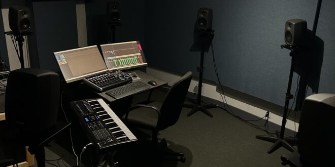 A darkly lit room with acoustic treatment on the walls, containing a desk with two computer monitors, a midi controller, and a keyboard and mouse on it. A desk chair is tucked under it. There are 6 speakers surrounding the desk.
