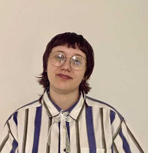 A person with short dark hair and glasses stands against a plain light-colored wall, wearing a striped button-up shirt with a string tie.