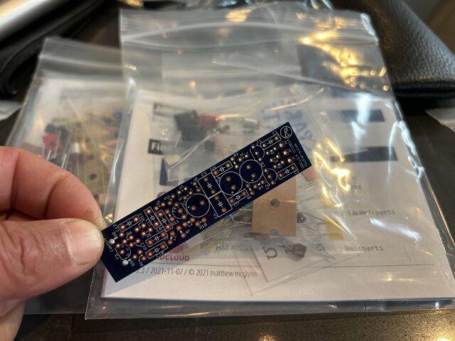 Eusebio's hand holding a small bare circuit board for a small condenser microphone, in front of a package of smaller parts