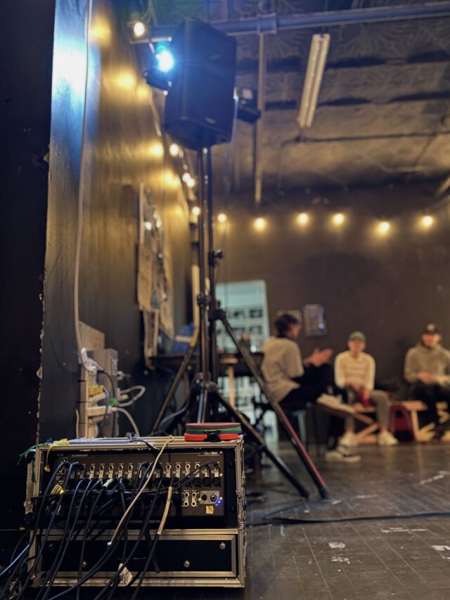 An Allen & Heath QU-SB sits on the floor with many cables hanging out of it. Above it is a QSC speaker on a stand. Blurred in the backgorund are Send + Recieve staff talking.