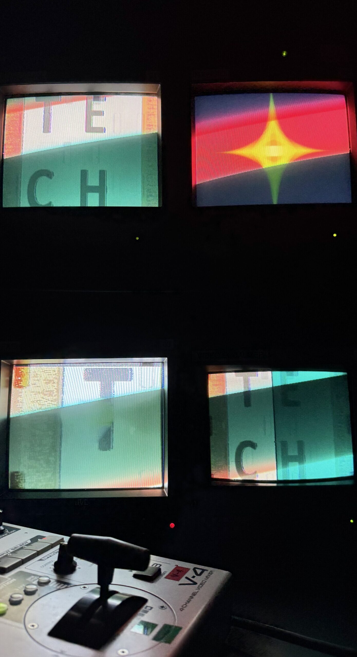 4 analog monitors display disorted images, two of which say TECH. There is an analog video mixer in the foregorund