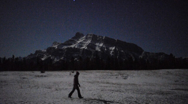 A person walks across a snowy plain at night with a mountain in the background and starry sky