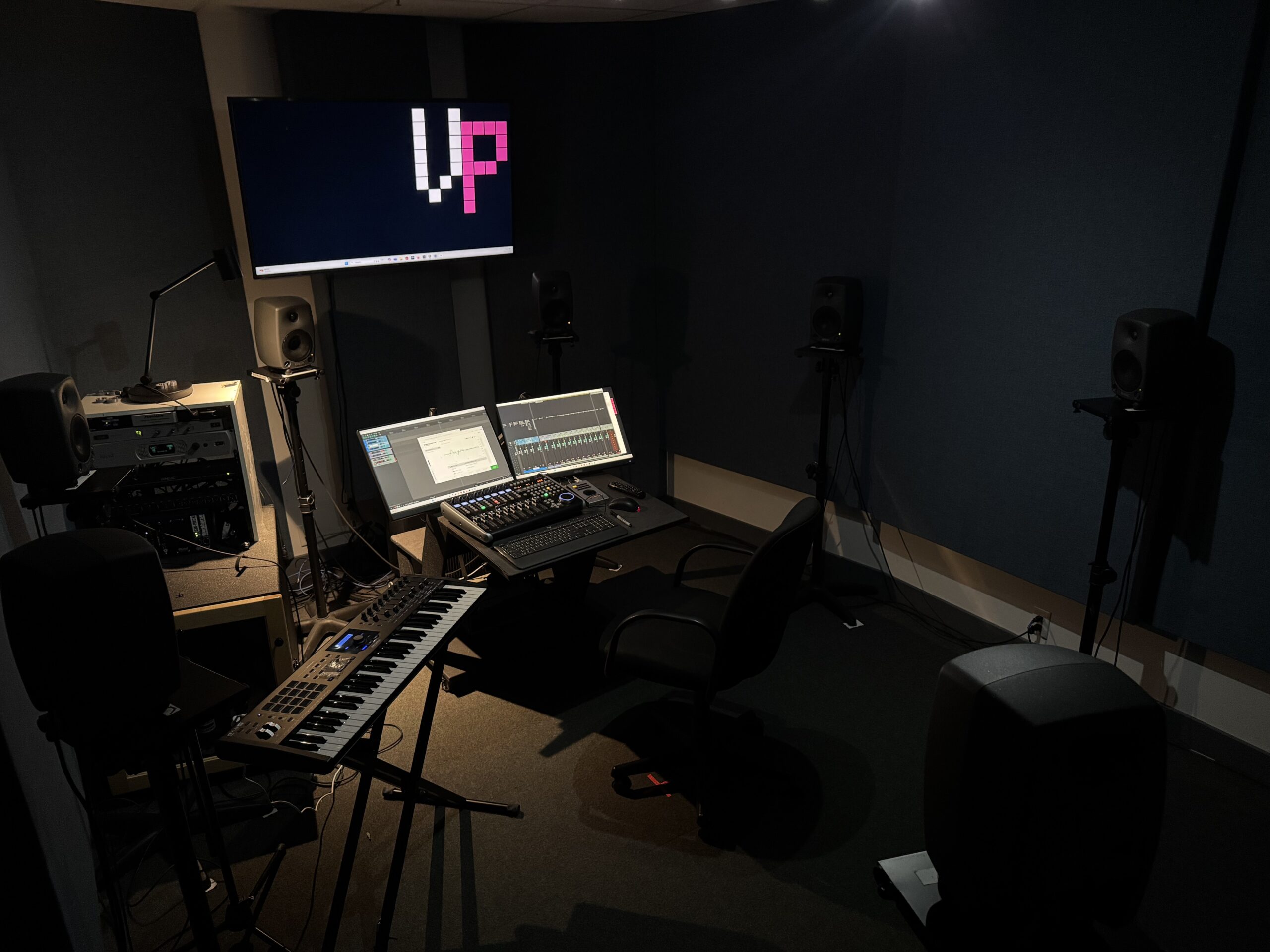 A darkly lit room with many speakers featured, and small desk with two monitors and a midi controller on it. To the side there is a midi keyboard