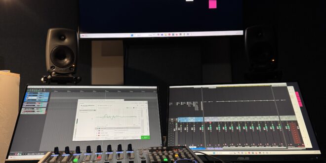 A dark audio suite with a large tv hung on the wall with the VP logo, underneath it a desk with tow monitors displaying recording software, and a midi controller with faders