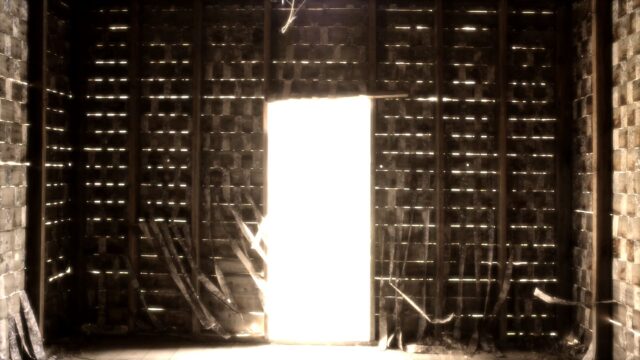 Inside wall of an old shack in darkness with a bright door way in the middle, light shines through the wood slats of the walls