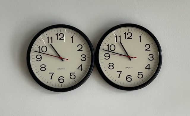 Two clocks on a white wall with their rims touching set to the exact same time