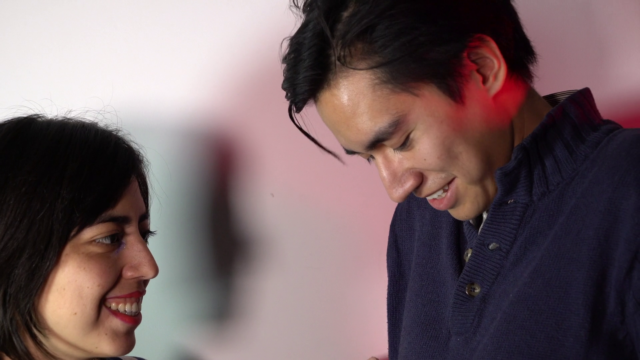 A man and woman smile at each other, the man looking at the ground shyly