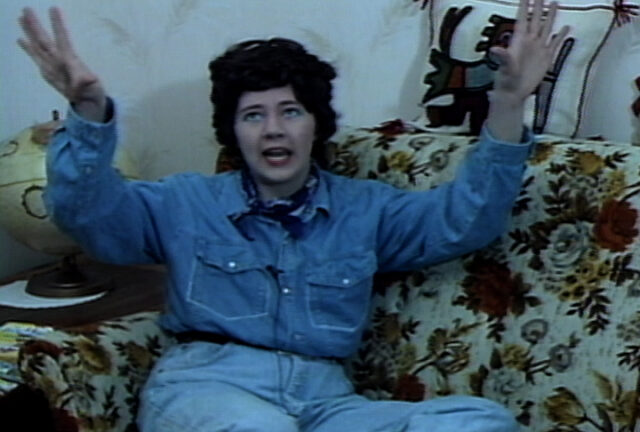 A woman on a vintage couch wearing denim-on-denim and a dark brunette wig with her hands expressively waving in the air