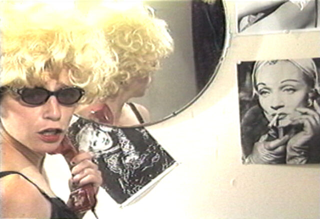 A woman looks into the camera in front of a mirror wearing a curly blonde wig and sunglasses with vintage photos pasted on the wall next to her