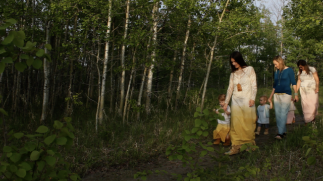 Three women walk through a wooded area holding hands with their toddlers