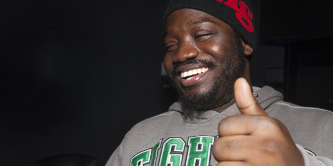 Iyunade laughing and wearing a black touque with red writing, giving a 'thumbs up' gesture