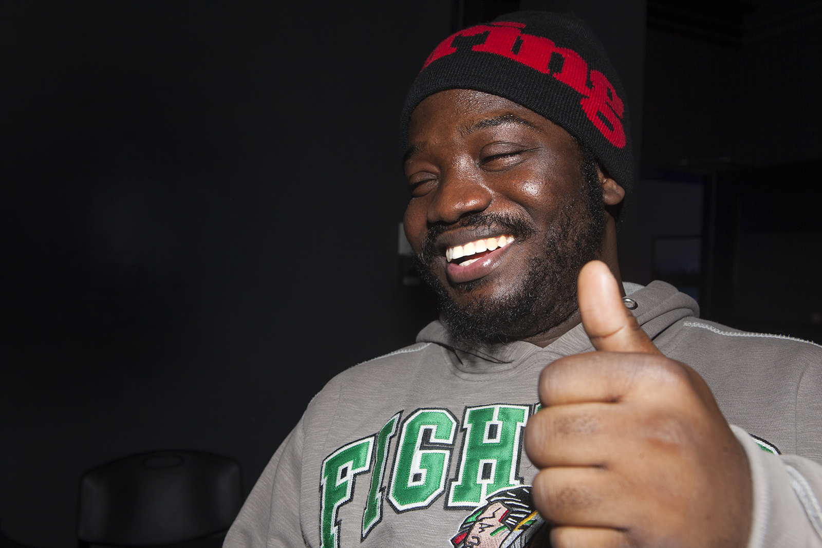 Iyunade laughing and wearing a black touque with red writing, giving a 'thumbs up' gesture