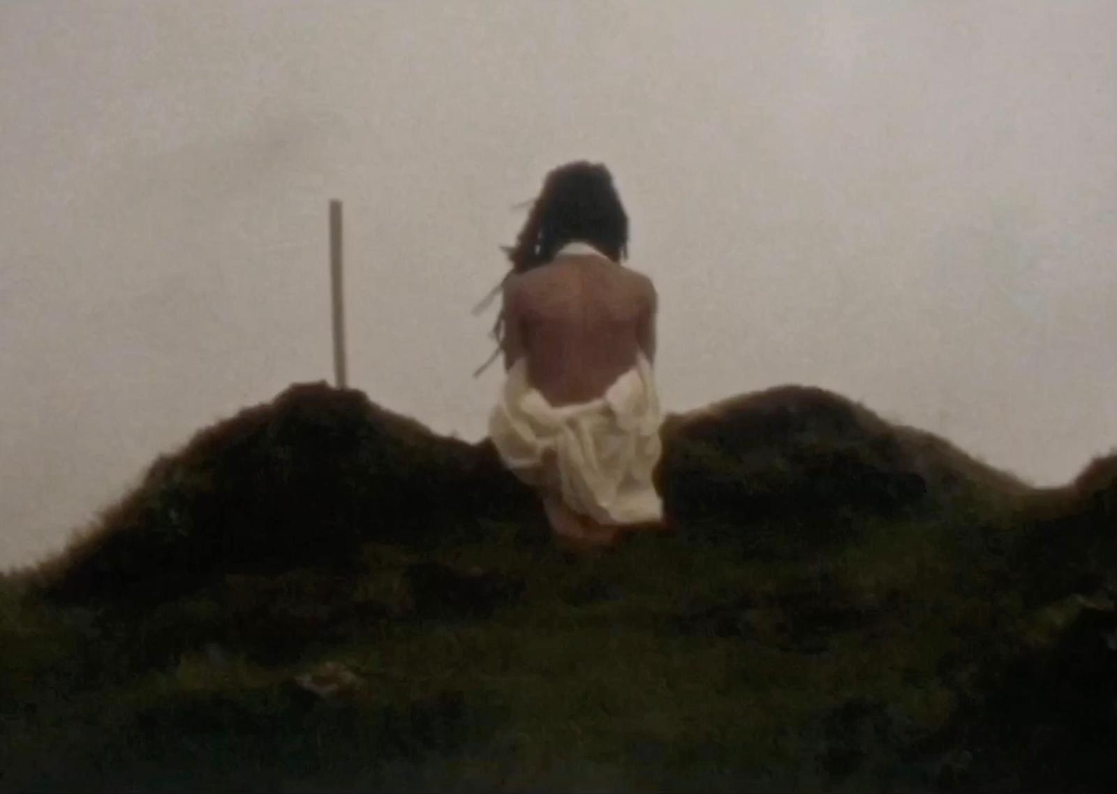 video still: a woman sits on a hill with her back to the audience. beyond her a grey cloudy sky sits ominously