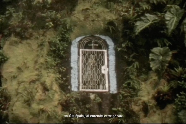 image still from video: a gate sits amongst blurry trees. the gate has a small cross above it.