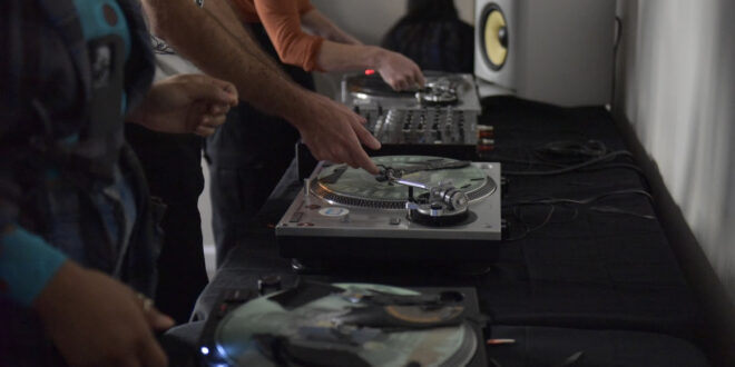 Side image of two turntables with two people standing in front, reaching toward the records