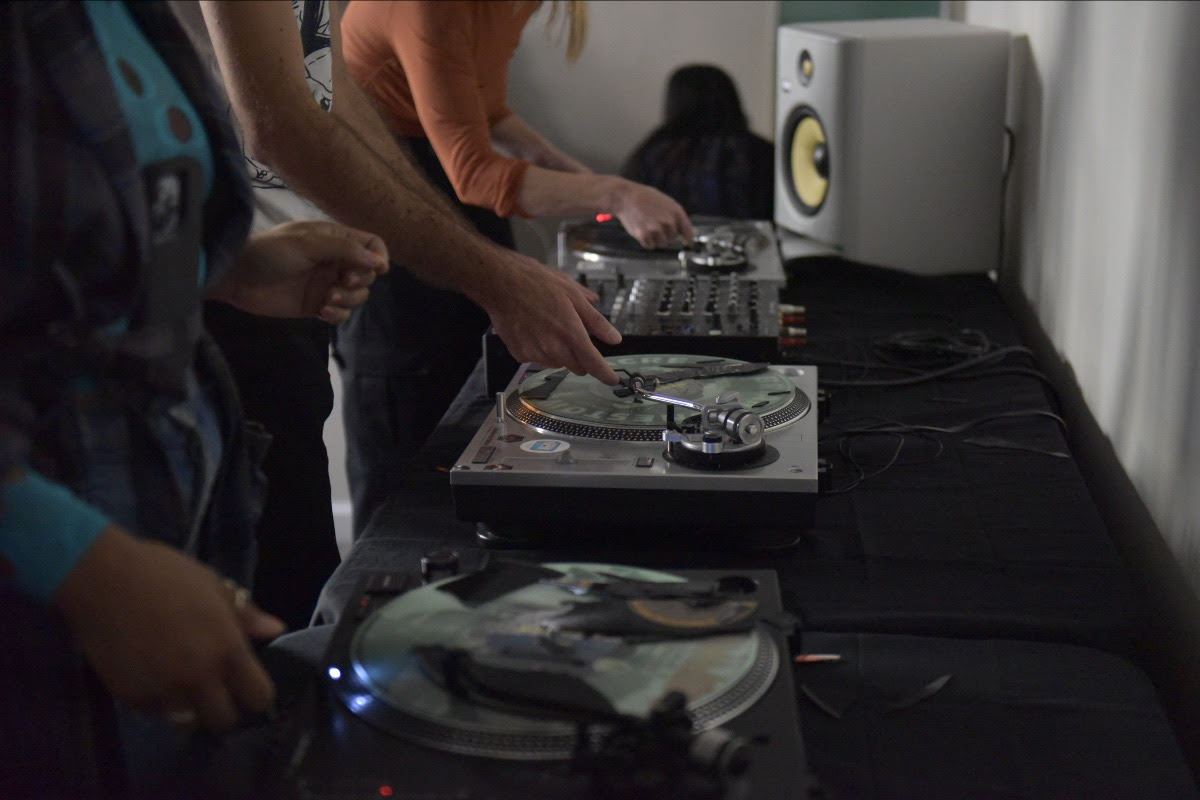 Side image of two turntables with two people standing in front, reaching toward the records